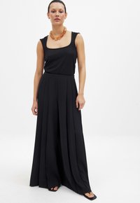 Zwarte mouwloze jumpsuit met een vierkante halslijn, vloeiende brede benen met plooien, en gecombineerd met een chunky amberkleurige ketting.