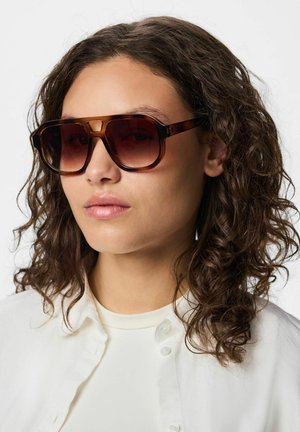 Woman with curly hair wearing dark brown oversized sunglasses and a white shirt against a neutral background.