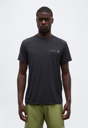 Mann steht frontal, trägt ein schwarzes kurzärmeliges T-Shirt mit "NORRØNA"-Logo und olivgrüne Shorts vor einfachem Hintergrund.