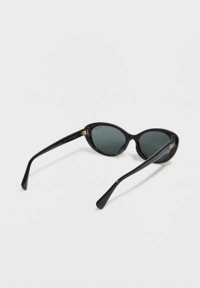 Μαύρα οβάλ γυαλιά ηλίου με σκούρα φακούς που είναι ανοιχτά και ακουμπούν σε ανοιχτή επιφάνεια, δείχνοντας το εσωτερικό των βραχιόνων και τις χρυσές λεπτομέρειες στις πτυχώσεις.