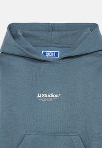 Φόρμα φούτερ με κουκούλα σε ανοιχτό μπλε ύφασμα, διαθέτει καγκουρό τσέπη, λευκό λογότυπο "JJ Studios" και ετικέτα με την αναγραφή "JACK & JONES."