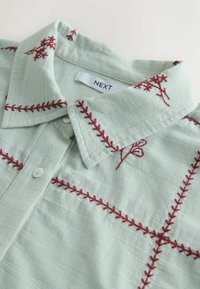 Chemise boutonnée vert clair avec des motifs floraux brodés rouges et des motifs de couture sur le col et la poitrine, avec une étiquette de marque "NEXT".