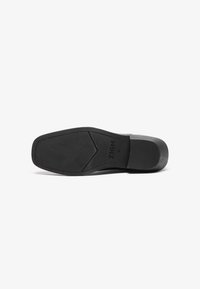 Suela de zapato negra con una superficie de goma texturizada, una forma angular con un ligero tacón y detalle en relieve con el nombre de la marca "ZIGN."