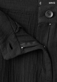 Tissu de pantalon noir en gros plan montrant un bouton, une fermeture éclair et une ceinture pliée avec une étiquette « Made in India » et le logo de la marque MNG.