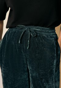 Samt-Hosen in einer dunklen Tealfarbe mit einem elastischen Bund, Kordelzug und Seitentaschen. Glatte Textur mit leichtem Glanz.