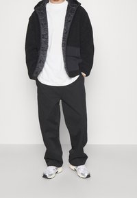 Schwarze Jacke mit strukturierter Oberfläche und satinartigem Finish, weißes T-Shirt, lockere schwarze Hose und graue Turnschuhe. Mit Kapuze und Taschen ausgestattet.