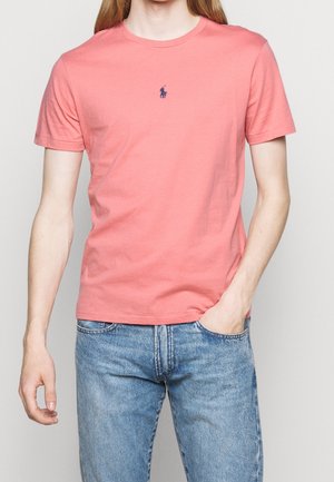 Koraliowa bawełniana koszulka z okrągłym dekoltem, krótkimi rękawami i małym granatowym logo na piersi. Noszona z jasnoniebieskimi dżinsami.