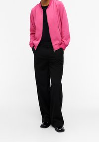 Giubbotto bomber rosa con polsini a coste e chiusura con zip, abbinato a pantaloni neri a gamba larga e scarpe nere lucide. Design semplice e moderno.