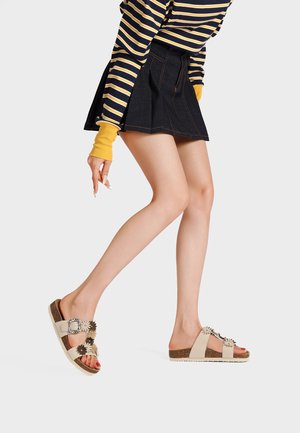 Donna che indossa sandali beige floreali con fibbia, gonna plissettata nera e maglia a righe manica lunga con polsini gialli, che cammina con un piede sollevato.