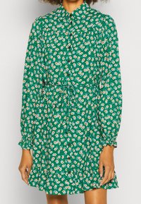 Groene bloemenjurk met lange mouwen, knoopsluiting aan de voorkant, strikceintuur, gerimpelde zoom, met kleine witte en oranje bloemen op een donkergroene achtergrond.