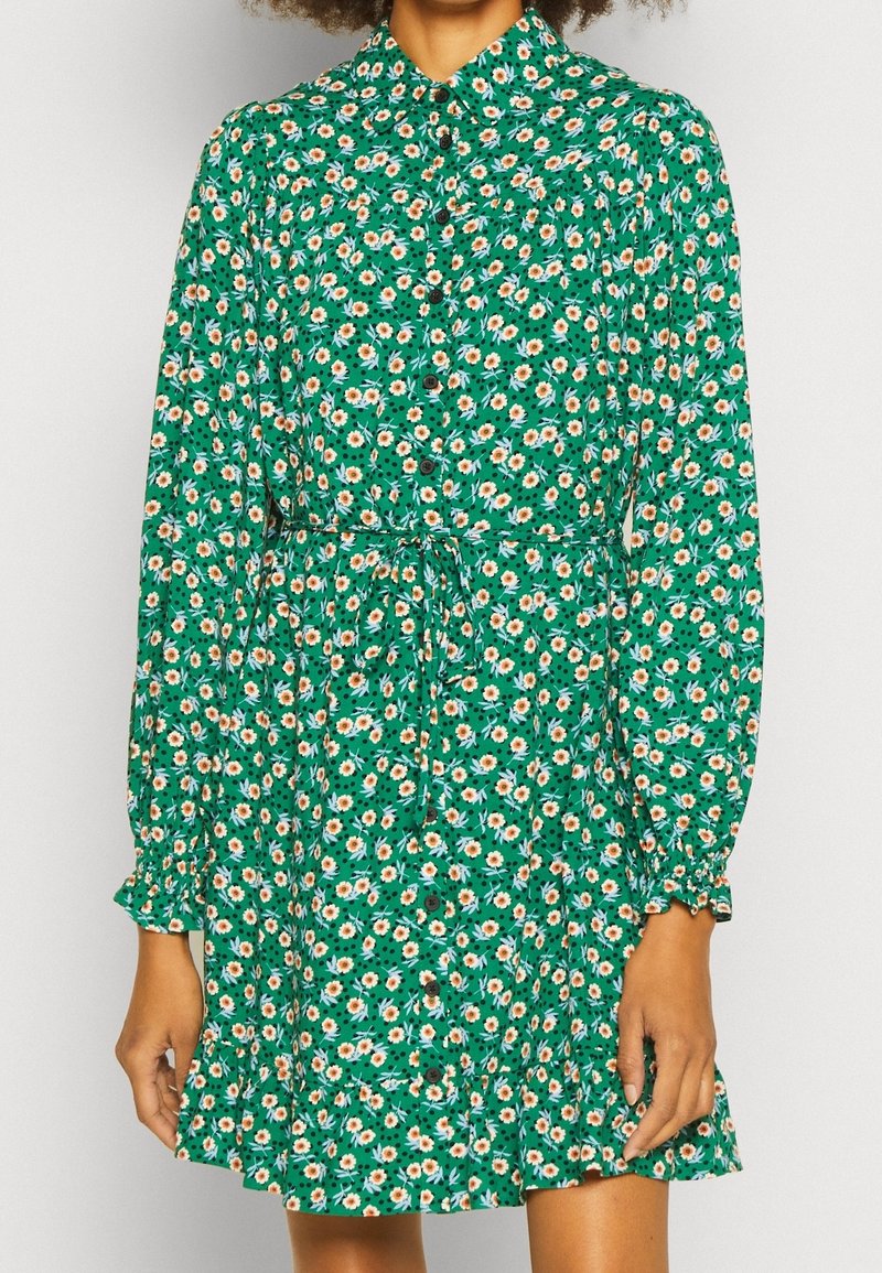 Groene bloemenjurk met lange mouwen, knoopsluiting aan de voorkant, strikceintuur, gerimpelde zoom, met kleine witte en oranje bloemen op een donkergroene achtergrond.