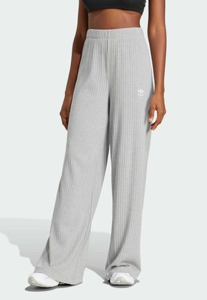 Femme portant un pantalon côtelé gris clair taille haute à jambes larges avec le logo Adidas et des baskets blanches, sur un fond clair uni.