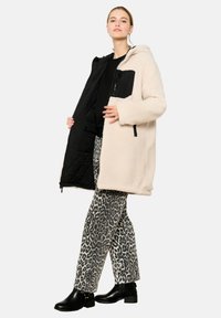 Béžová sherpa bunda s čiernou zipsovou vreckom, kapucňou a čiernou podšívkou; skombinovaná so sivými nohavicami s leopardím vzorom a čiernymi členkovými bootami.