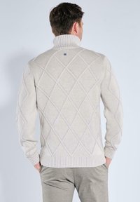 Cremfarbener Strickpullover mit hohem Kragen, der ein Rautenmuster aufweist, sowie gerippten Bündchen und Saum. Auffälliges Markenlogo auf der Rückseite.