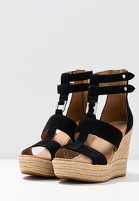 Czarne zamszowe sandały na koturnie z otwartym noskiem, paskami na kostkę i plecioną jucianą podeszwą. Zawierają srebrne akcesoria i miękkie beżowe wnętrze.