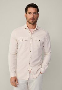 Camisa de manga larga color beige claro con dos bolsillos frontales, cierre de botones y una textura suave. Llevada sobre una camiseta de rayas rosas.