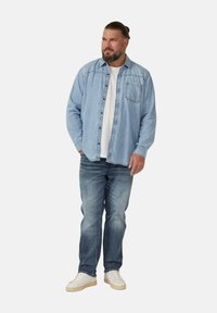 Man met een baard en teruggebonden haar, gekleed in een lichtdenim shirt, een wit T-shirt, vervaagde blauwe jeans en witte sneakers, staat met een hand in zijn zak.
