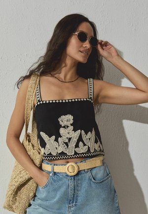 Žena s kulatými slunečními brýlemi, černým vyšívaným krátkým topem, džínami s tkaným páskem a velkou tkanou kabelkou přes rameno.