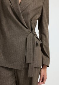 Ruskea rakennekas blazer, jossa on sivusoljentaa, yhdistettynä vastaaviin housuihin, korostaen puvulle omistettua istuvuutta ja kankaan kuvioita.
