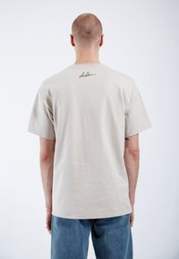 Beige bavlněné tričko, krátké rukávy, volný střih, s malým podpisem grafiky na horní části zad. Nošeno s modrými denimovými džíny.