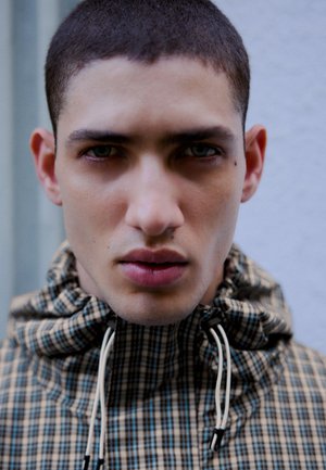 Mladý muž s krátkymi vlasmi, oblečený v károvanom vetrovke s vysokým golierom a šnúrkami, ktorý hľadí priamo do objektívu na jednoduchom pozadí.