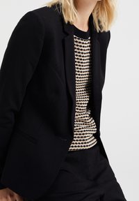 Svart kavaj med smal passform och notch lapel; bärs över en strukturerad beige och svart m önstrad tröja, matchad med byxor i samma stil.