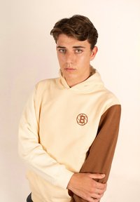 Sudadera con capucha que presenta un cuerpo beige y una manga marrón. Tejido suave, logotipo bordado en el pecho y puños acanalados.