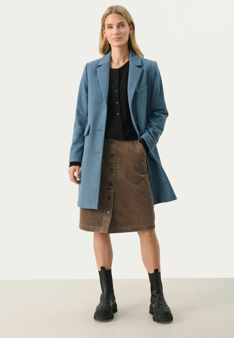 Jasnoniebieski płaszcz z wełny, czarny sweter na guziki, brązowa spódnica dżinsowa z guzikami z przodu oraz masywne czarne botki na kostkę.