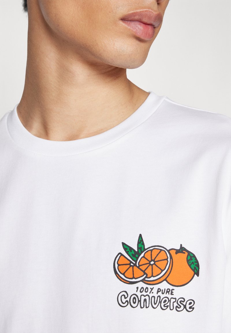 Camiseta blanca de manga corta, con un gráfico colorido de fruta naranja y el texto "100% PURO CONVERSE" en el pecho.