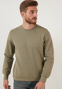 Sudadera de manga larga en verde olivo, confeccionada en tejido suave, con cuello redondo, puños acanalados y una superficie lisa y texturizada.