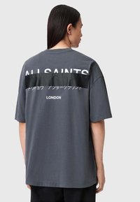Šedé bavlněné tričko s velkým bílým textem na zadní straně, který zní "ALLSAINTS" a "LONDÝN", s lesklým černým pruhem uprostřed.
