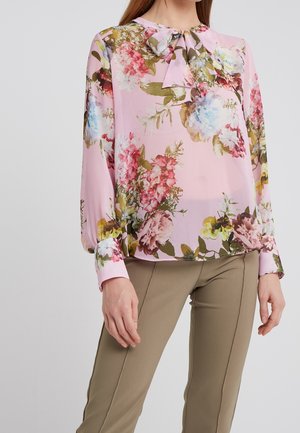 Frau trägt eine rosa Bluse mit Blumenmuster, Schleifenkragen und langen Ärmeln, kombiniert mit beige geschnittenen Hosen.