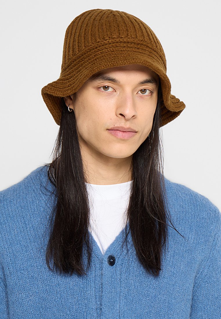 Brauner gerippter Bucket Hat aus strukturiertem Material, mit breiter Krempe und flexibler Form, getragen mit einem blauen Cardigan über einem weißen Hemd.