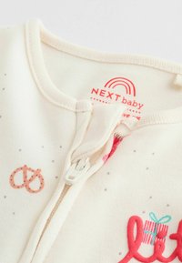 Cremefarbener Baby-Overall mit Reißverschluss, mit rotem und türkisfarbenem Text. Mit grafischen Darstellungen eines pinken Brezels und Geschenks. Weicher Stoff mit kleinen grauen Punkten.