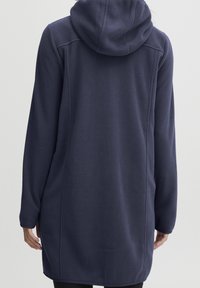 Chaqueta de forro polar azul marino larga con capucha, que presenta una textura suave, costuras laterales y un corte recto, llegando hasta la mitad del muslo.