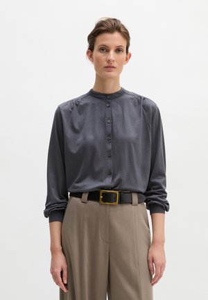 Vrouw die een donkergrijze blouse met knopen draagt, met gerimpelde mouwen, gestopt in een beige broek met hoge taille, vastgemaakt met een zwarte riem met een gouden gesp.