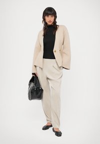 Beige op maat gemaakte blazer met twee gouden knopen, zwarte coltrui, licht gestreepte hoge taille broek en zwarte draagtas. Platte zwarte schoenen.