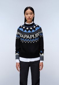 Czarny sweter dzianinowy z niebieskimi i białymi wzorami geometrycznymi, okrągłym dekoltem i kontrastującymi szarymi rękawami. Noszony na białej koszuli z kołnierzykiem.