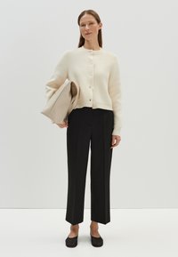 Crème-kleurige cropped cardigan met vijf knopen, gecombineerd met zwarte rechte broek en zwarte platte schoenen. De tas is van een glad, beige materiaal.
