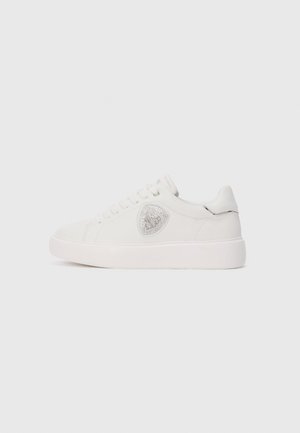 Sneaker basse blanche avec lacets, arborant un emblème argenté sur le côté et un détail argenté discret sur l'onglet du talon.