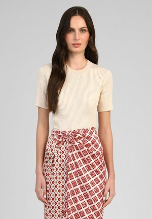 Femme aux longs cheveux foncés portant un haut court en maille côtelée crème et une jupe portefeuille rouge et blanche à motifs, nouée à la taille.