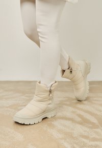 Bottines molletonnées beiges avec fermeture éclair, présentant une semelle texturée et un design épais et imposant. Matériau doux pour un confort optimal.