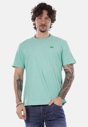 Giovane uomo con capelli ricci, indossa una maglietta verde chiaro e jeans blu, mostra tatuaggi dettagliati su entrambi gli avambracci, in piedi contro uno sfondo bianco.