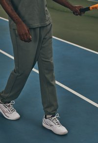 Sive atletske trenirke z elastičnimi manžetami, v kombinaciji z belimi superge, ki imajo sive akcentne barve. Tenis raketa v roki. Notranje igrišče v ozadju.