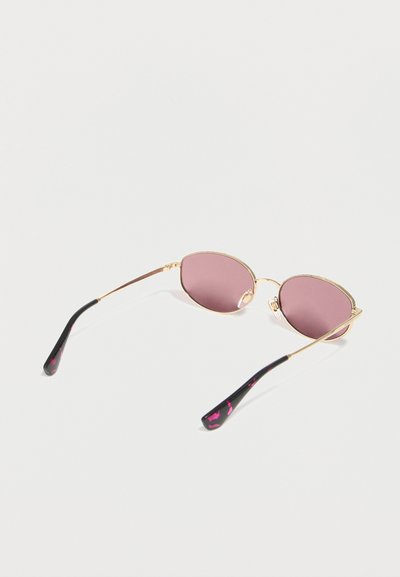 Γυαλιά ηλίου με χρυσή κορνίζα και οβάλ ροζ φακούς, με μαύρα σκέλη που έχουν ροζ λεπτομέρειες, ακουμπισμένα σε λευκή επιφάνεια.