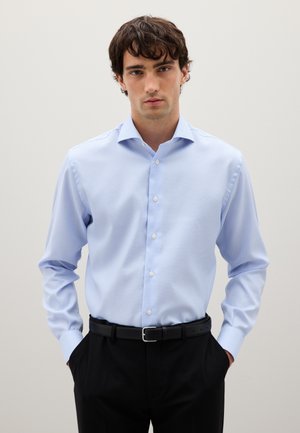 Giovane con i capelli ricci e scuri indossa una camicia a quadri azzurri chiari infilata in pantaloni neri con una cintura nera, in piedi con le mani in tasca.