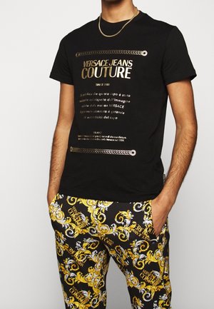 Uomo che indossa una t-shirt nera Versace Jeans Couture con scritta dorata e pantaloni neri con stampa barocca ornata gialla e bianca.