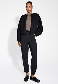 Giacca nera a cropped con maniche ampie, maglione marrone strutturato, pantaloni neri con vita elasticizzata e scarpe nere stringate. Design minimalista.