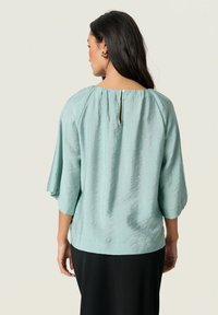Blusa azzurro chiaro a maniche lunghe con una texture morbida e un dettaglio a occhiello sulla schiena. Presenta una vestibilità ampia e maniche larghe, abbinata a un pantalone nero.