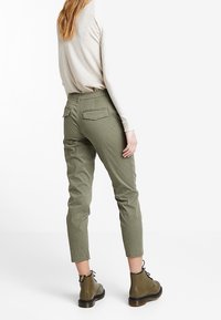 Olive zielone zwężane spodnie z dwoma tylnymi kieszeniami, zestawione z brązowymi sznurowanymi butami. Materiał wydaje się mieć strukturę o swobodnym kroju.